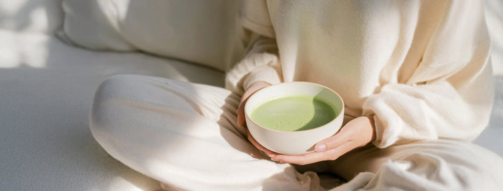 A person in a cozy cream lounge set holds a bowl of green probiotic matcha, embodying a calm morning wellness ritual.