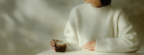 Person in a white sweater holding a glass of SUPERBA COFFEE at a sunlit table, calm and mindful coffee ritual