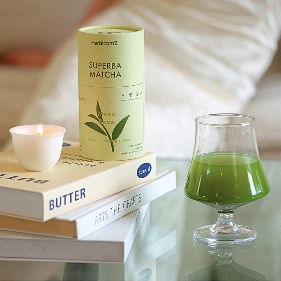 HerbloomZ SUPERBA MATCHA tin with warm green cup and candle on books — probiotic matcha, low-acid, non-fizzy, gut-friendly ritual