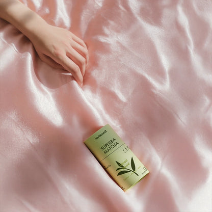 HerbloomZ SUPERBA MATCHA tin on blush silk beside a relaxed hand — probiotic matcha, gut-friendly, low-acid warm ritual
