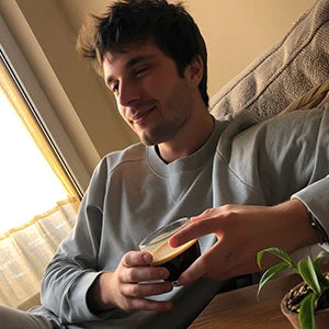 Young man sitting on a sofa in morning light, holding a clear glass cup of SUPERBA Coffee and smiling  — crash-free energy and focus.